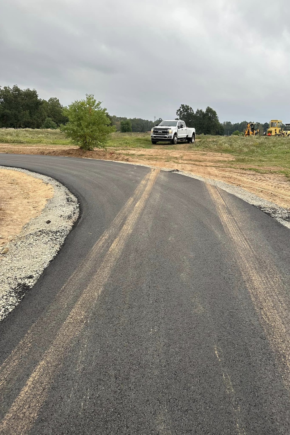 Paved driveway with a curve and a split, leading to a white truck parked in a field. Cloudy sky.