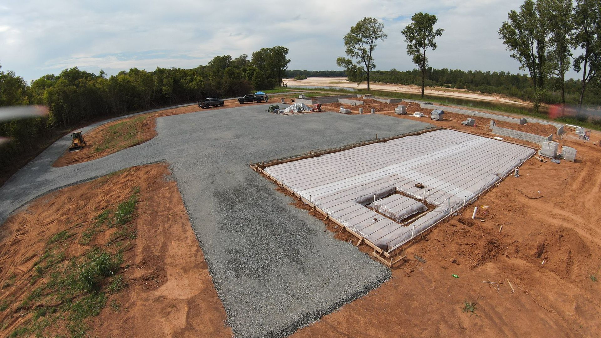 Construction site with gravel driveway, prepared concrete foundation, and surrounding dirt.