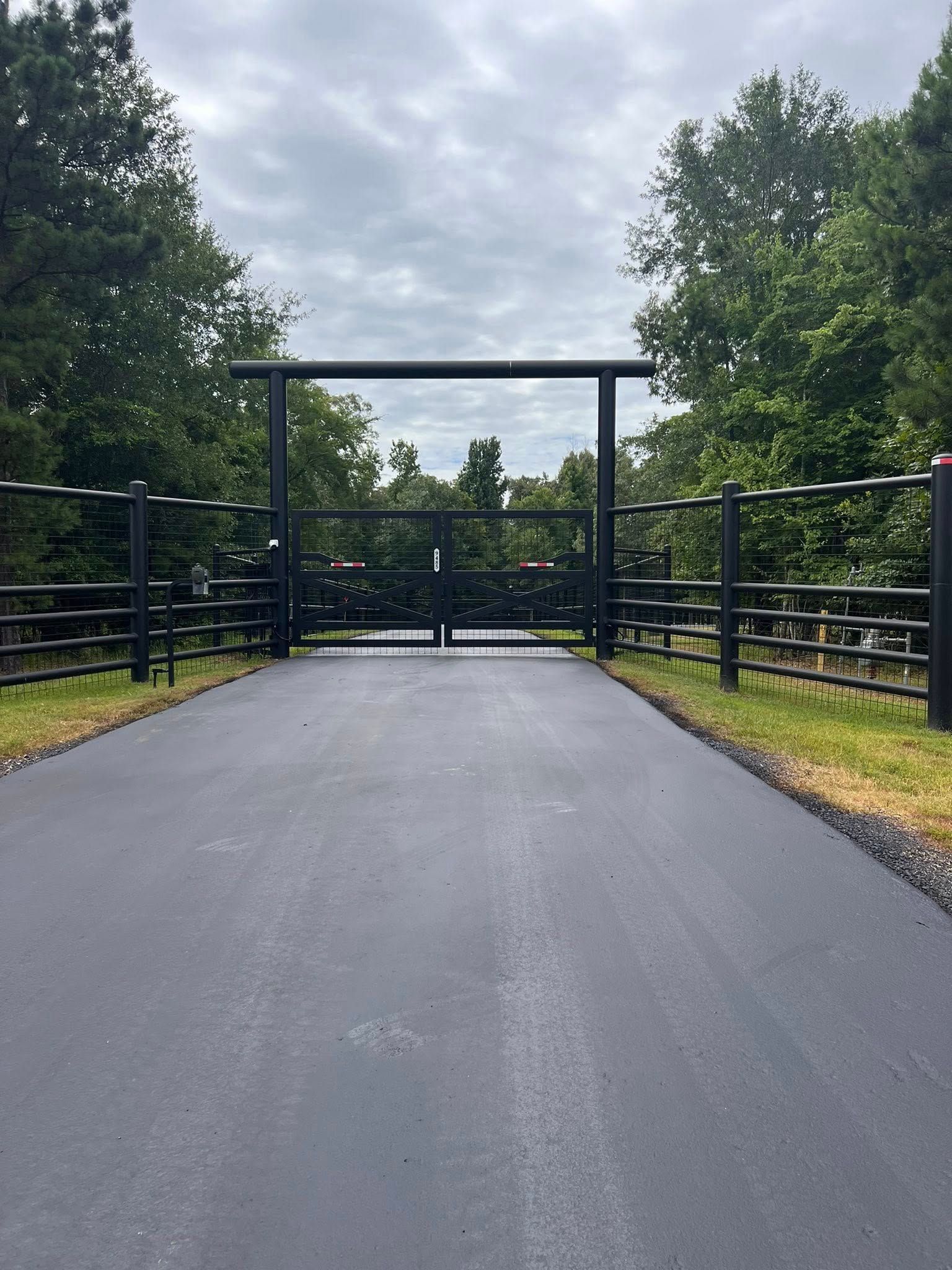 Black metal gate on asphalt driveway, flanked by matching fencing; trees in the background, overcast sky.