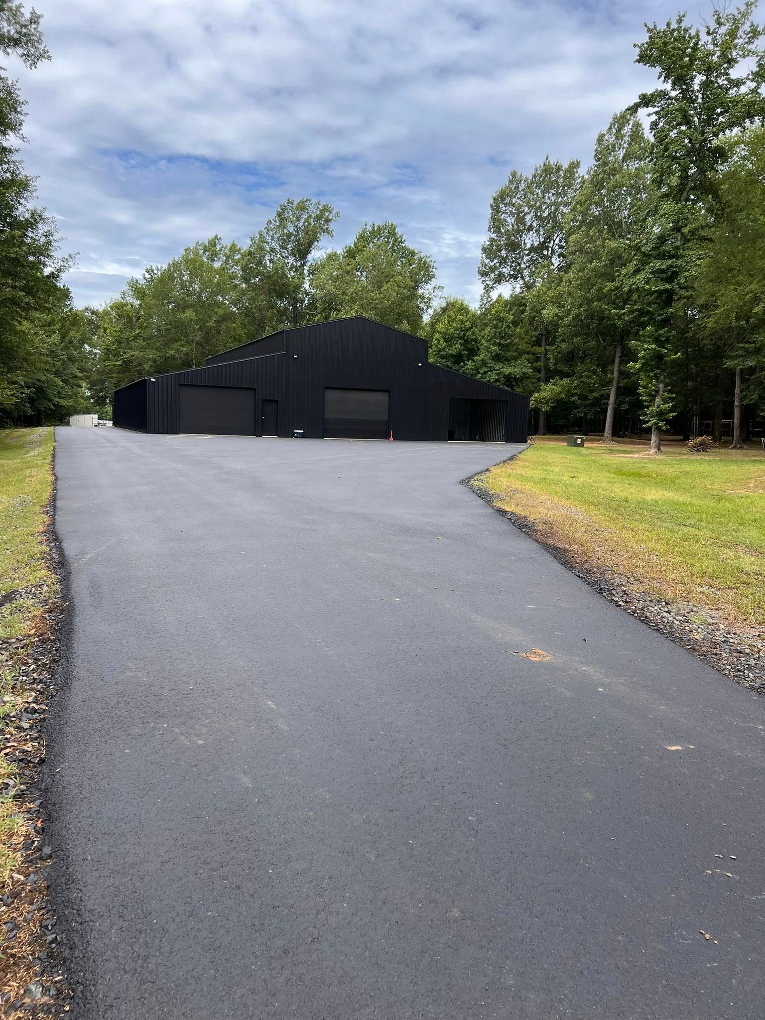 Black building with two garage doors and a paved driveway surrounded by trees.