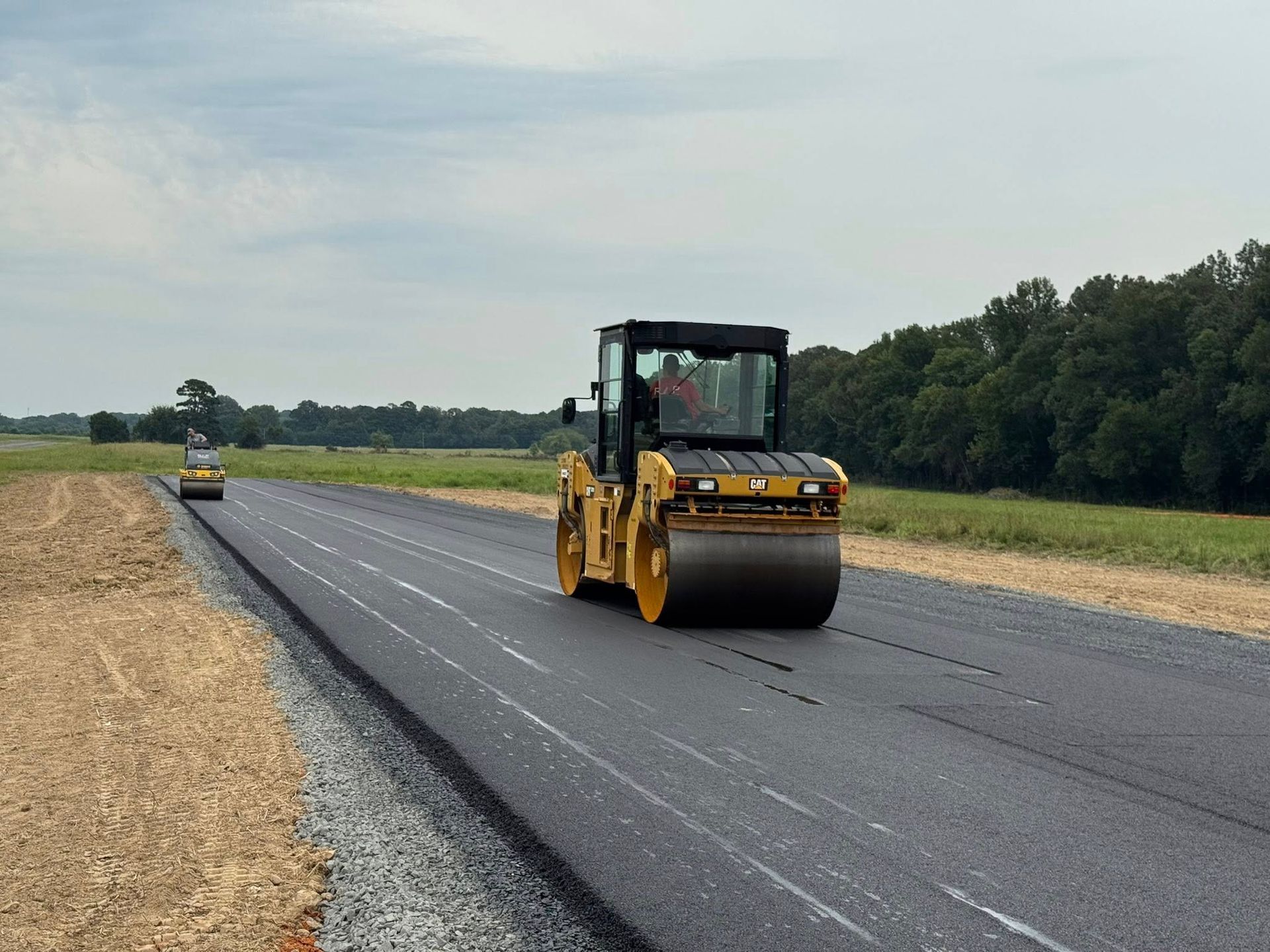 Yellow road rollers compacting fresh asphalt on a new road under a cloudy sky.