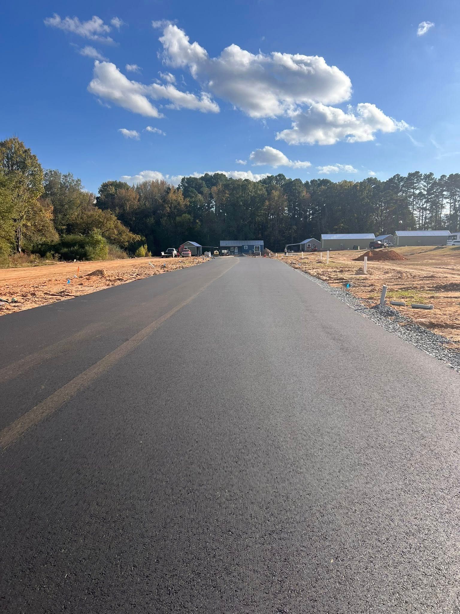 Newly paved road leading towards buildings, trees, and blue sky with clouds.