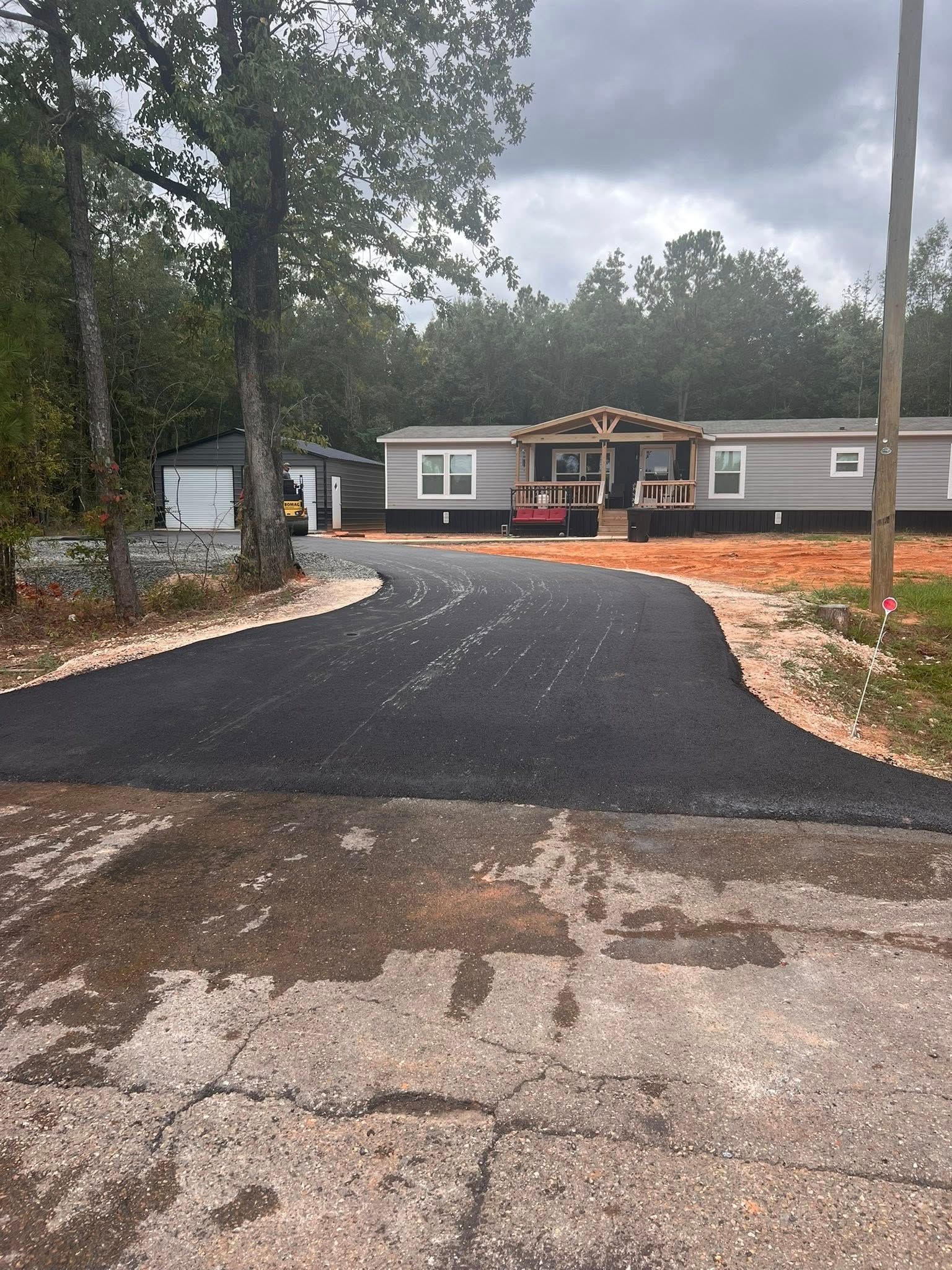 Black asphalt driveway leading to a gray manufactured home with a porch, shed to the left.