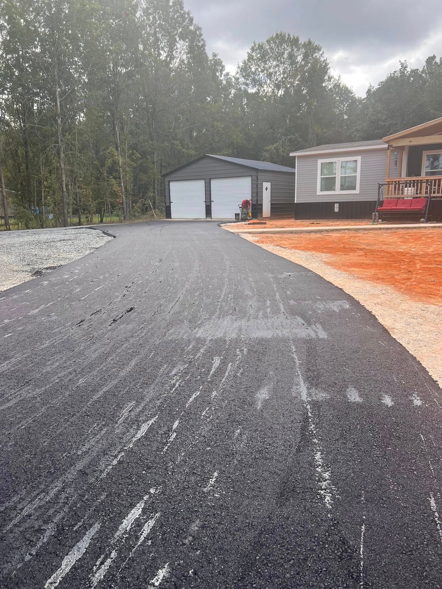 Asphalt driveway leading to a garage and a house, trees in the background. Overcast day.