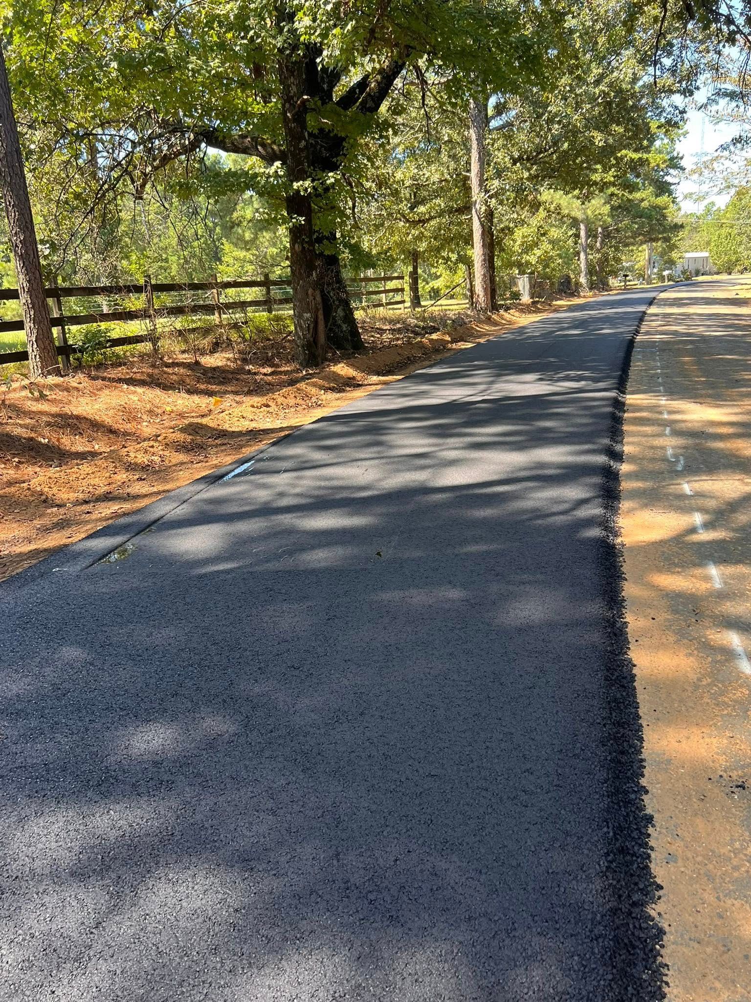 Newly paved asphalt road through a wooded area. The road is dark grey with yellow and brown shoulders.