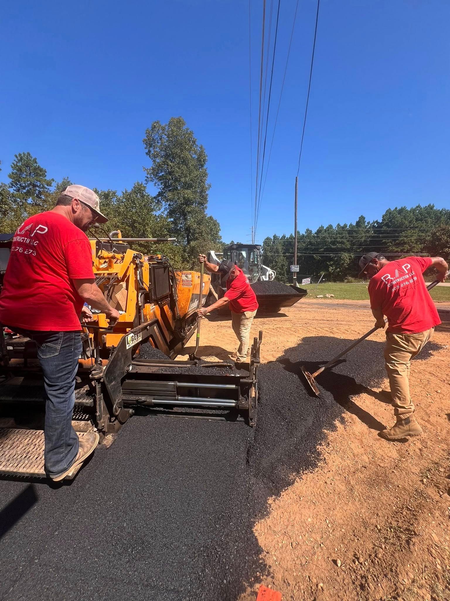 Construction workers paving a road on a sunny day. One operates a paving machine, two shovel asphalt.