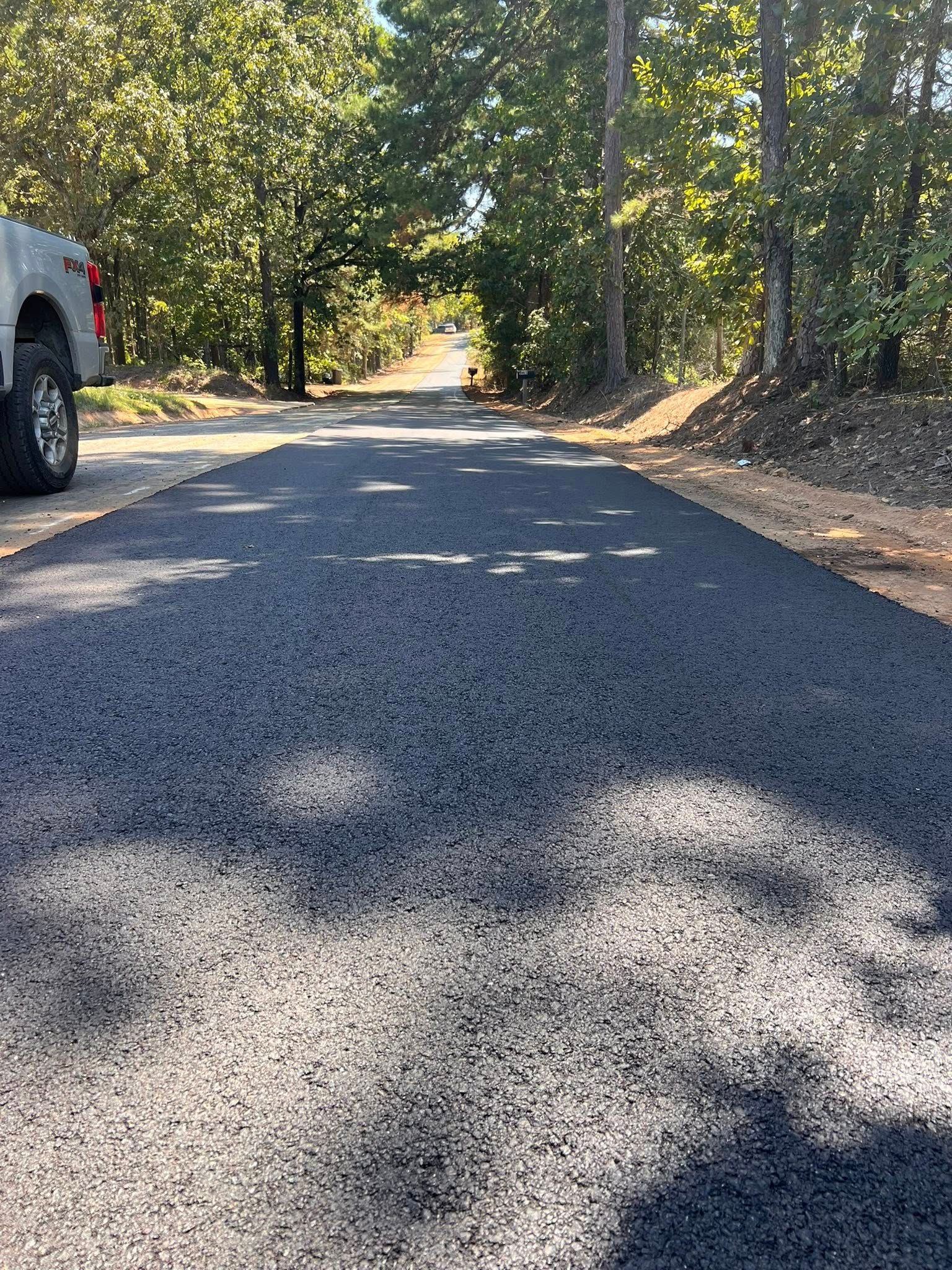 Newly paved asphalt driveway leading into a tree-lined area; a truck is parked on the left.
