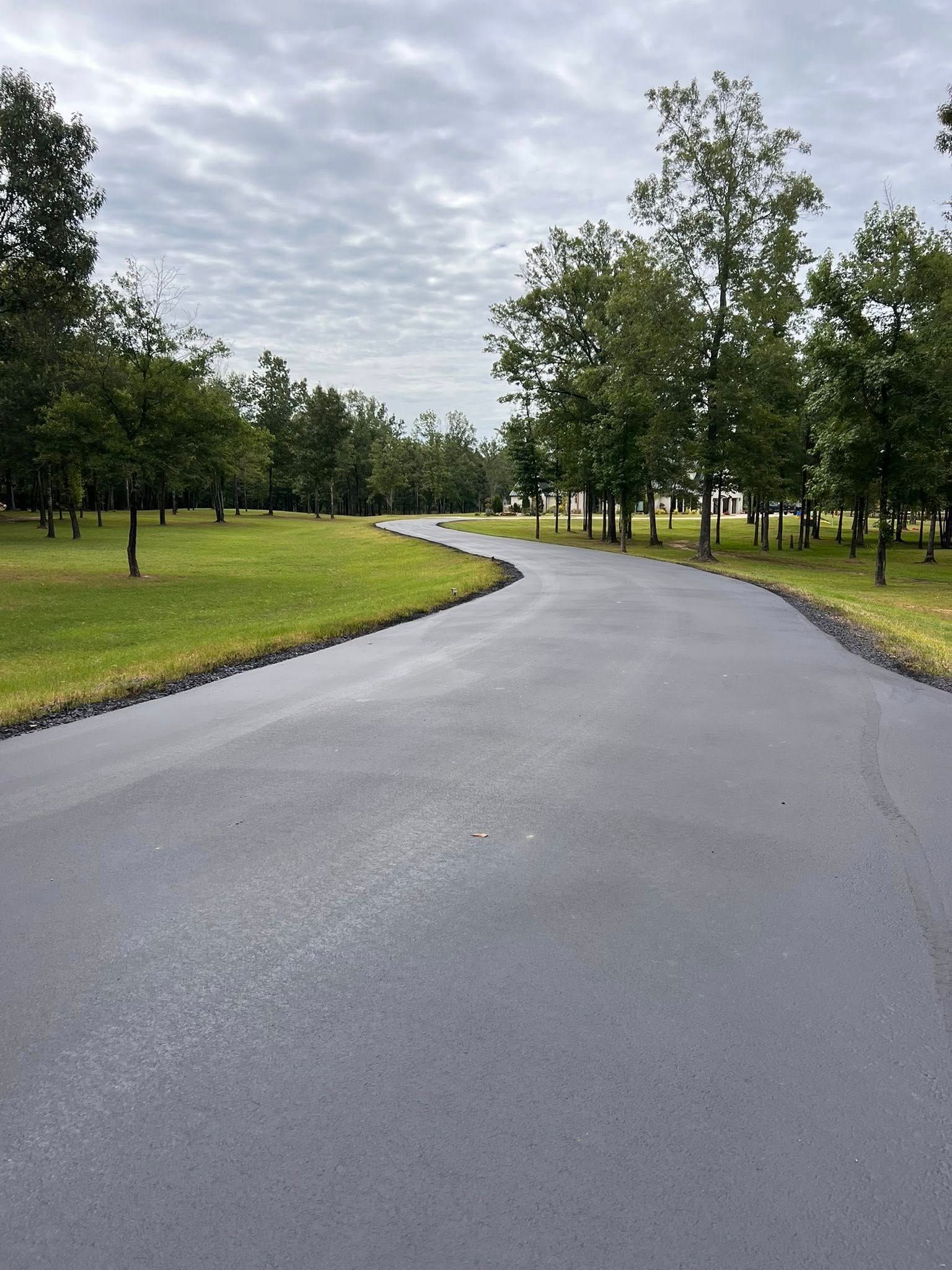 Paved road curves through a green grassy area with trees under an overcast sky.
