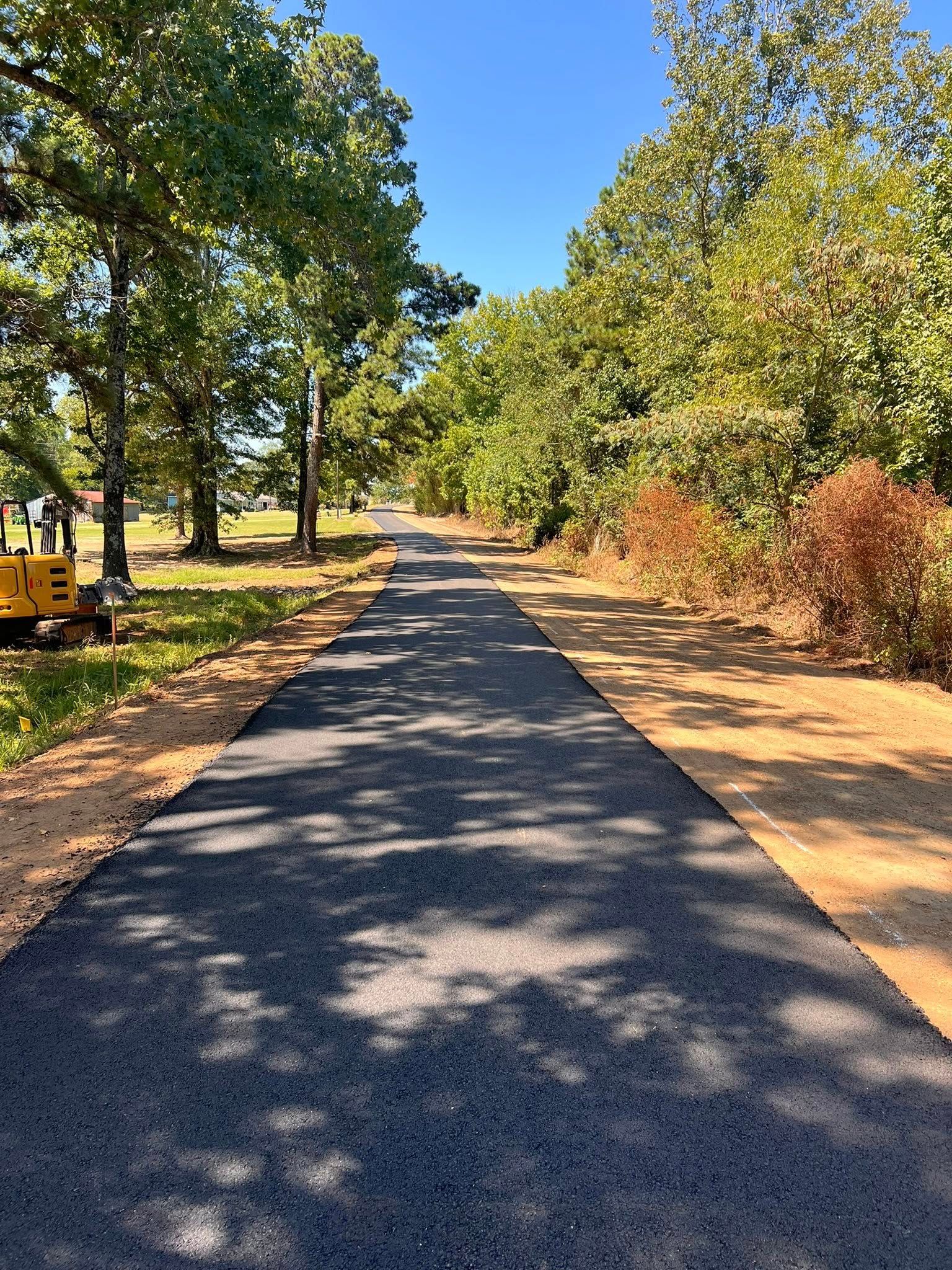 Newly paved asphalt path lined with trees and dirt shoulders under a bright blue sky.