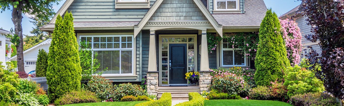 A craftsman-style house with blue-grey siding, a covered porch, and lush green landscaping on a sunny day.