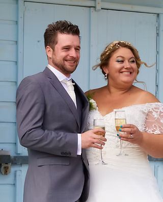 Smiling couple in wedding attire holding drinks, standing in front of a light blue door.
