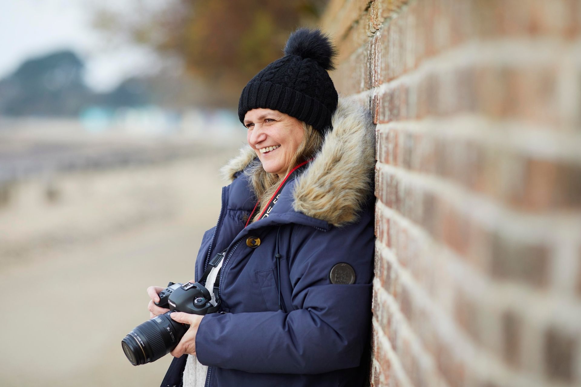 Person in a black hat and blue coat holding a camera, leaning against a brick wall outdoors on a chilly day