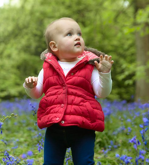 Toddler in a red vest standing in a flower-filled forest, looking up with hands raised