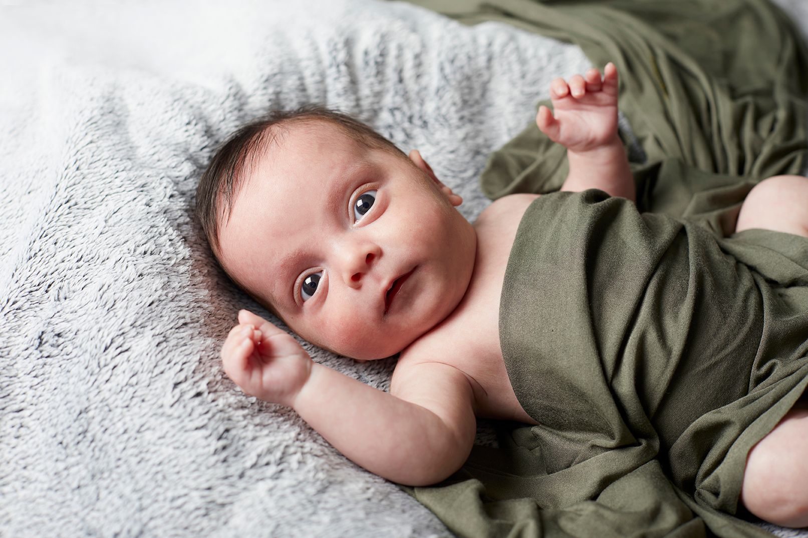 Baby lying on a fuzzy blanket, wrapped in green cloth, with hands raised and eyes open