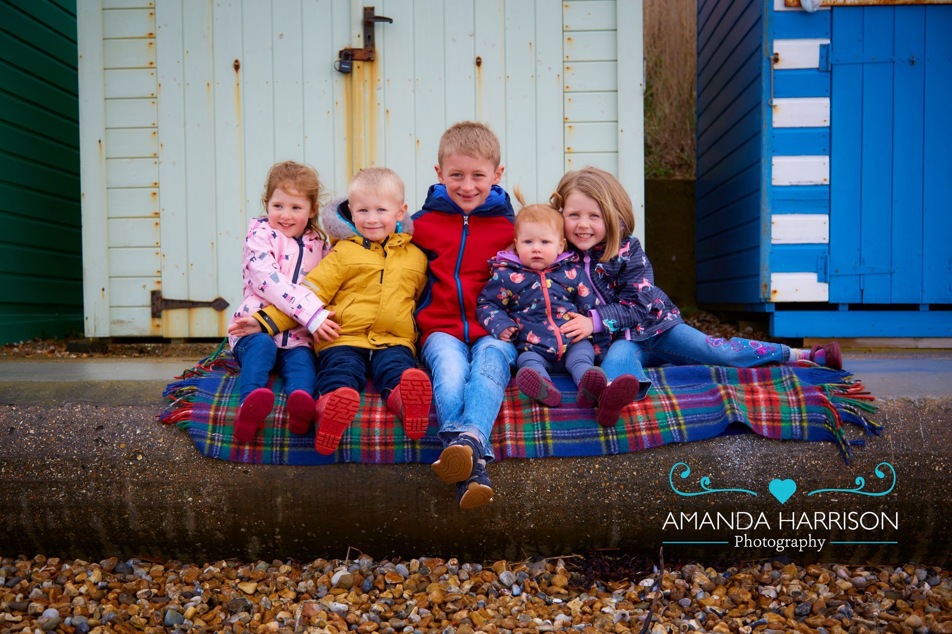 Four children sitting on a plaid blanket in front of a weathered house, smiling outdoors.