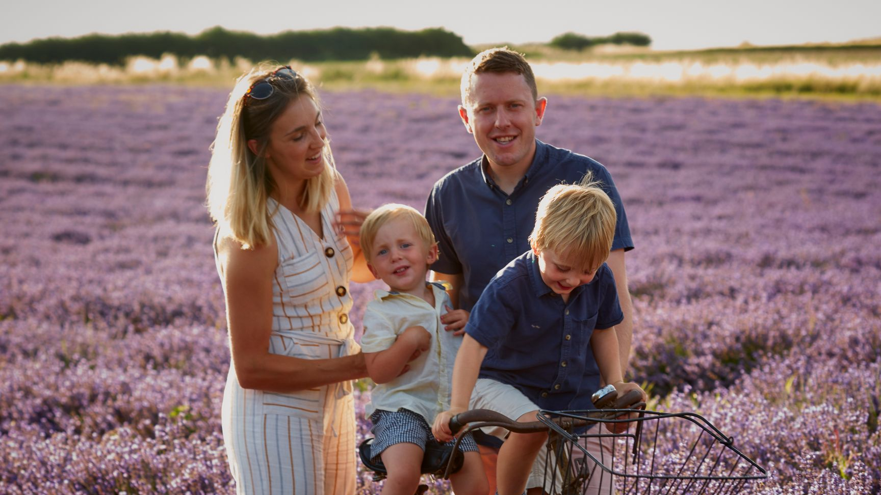 Family standing in a purple flower field, smiling beside a bicycle at sunset