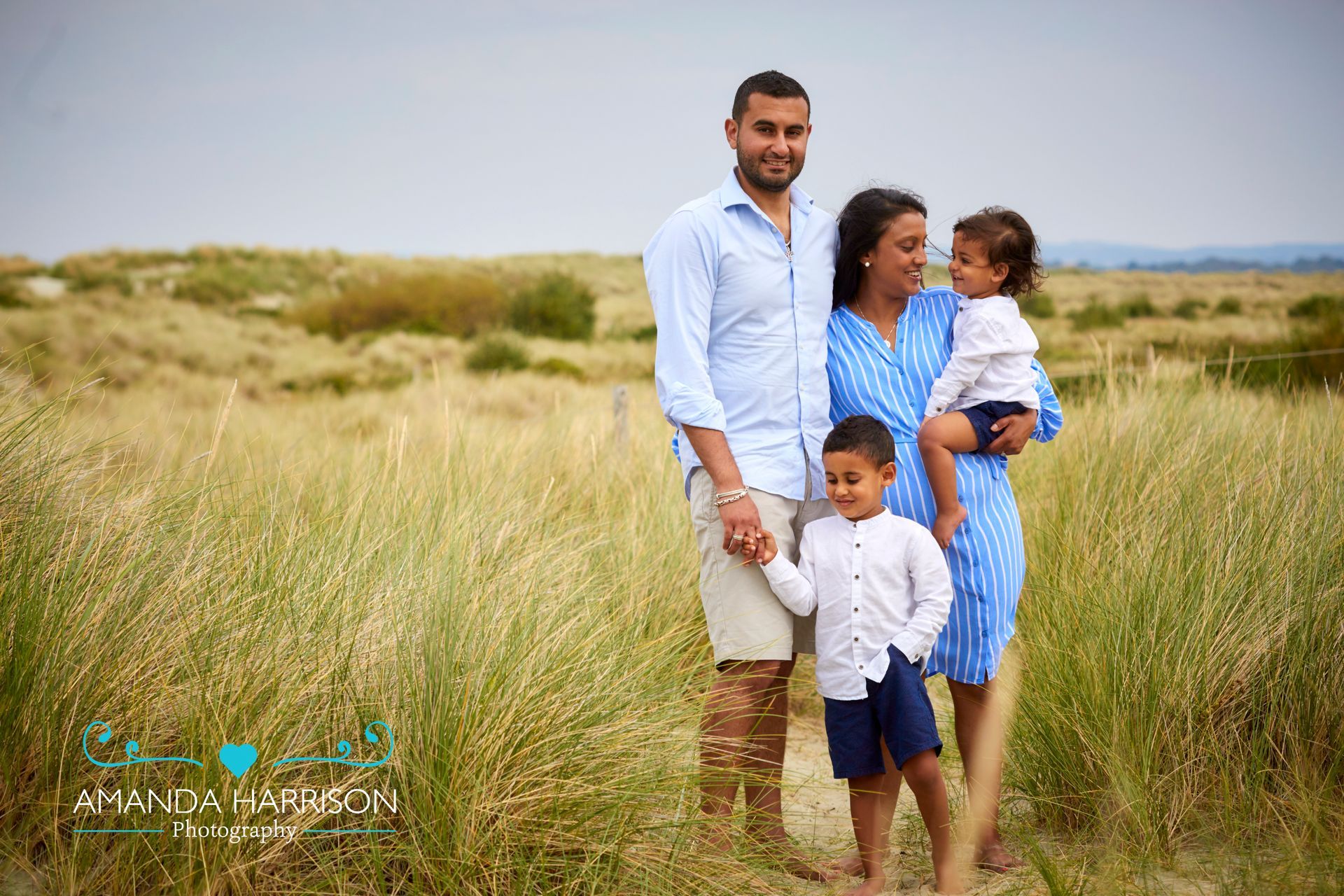 Family of four standing in tall grass on a beach, with a man, woman, and two young children.