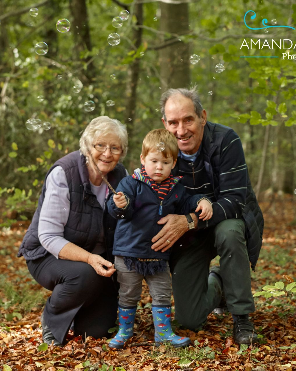 Three people crouch in a wooded area, holding a toddler outdoors in autumn leaves.
