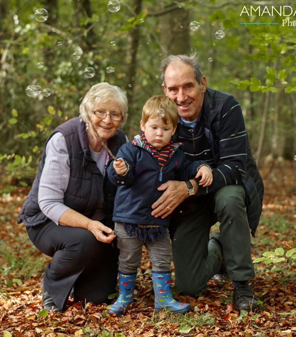 Three people crouch in a wooded park, smiling among fallen autumn leaves.