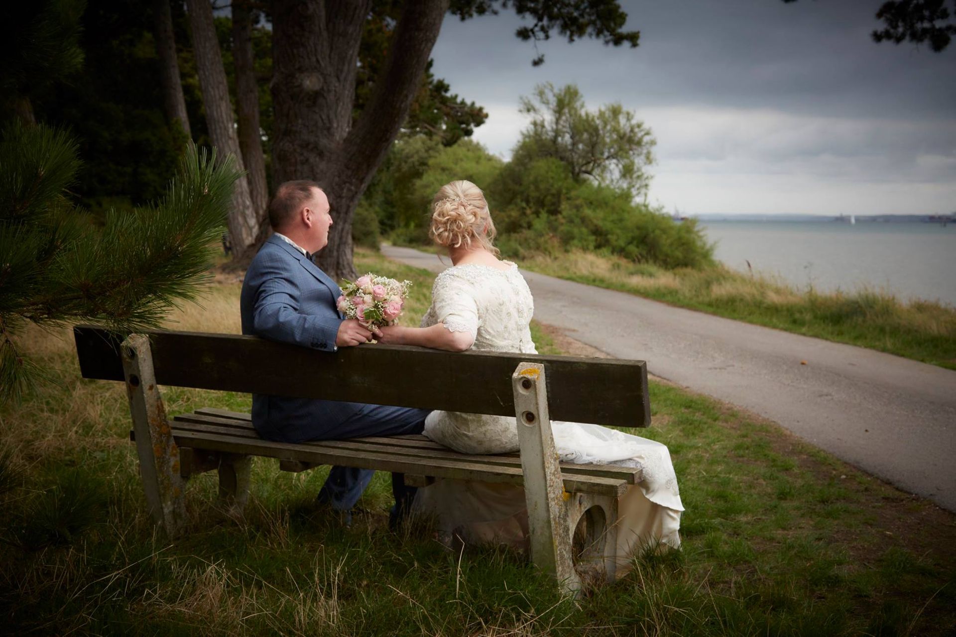 Couple holding hands on a bench by a coastal path with trees and water in the background