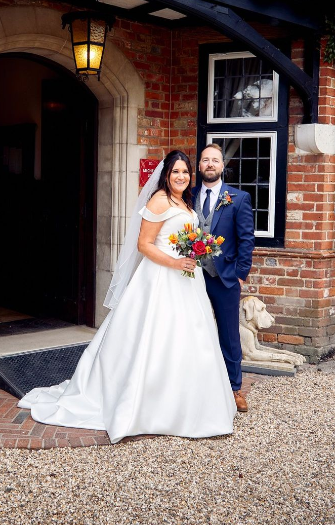 Bride and groom posing outside a brick building, bride holding a colorful bouquet and smiling