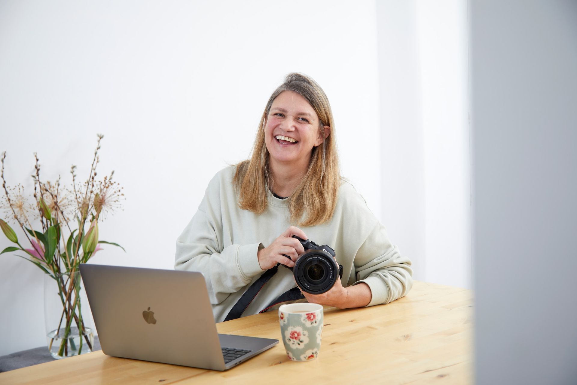 Smiling woman holding a camera at a wooden table with a laptop, mug, and vase of flowers