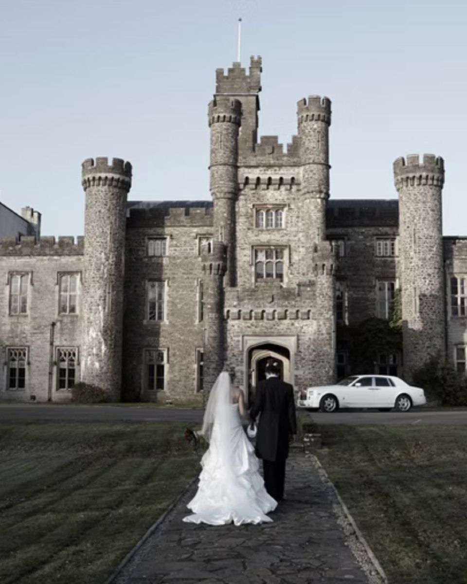 Bride and groom walking toward a stone castle entrance, with a white car parked nearby.