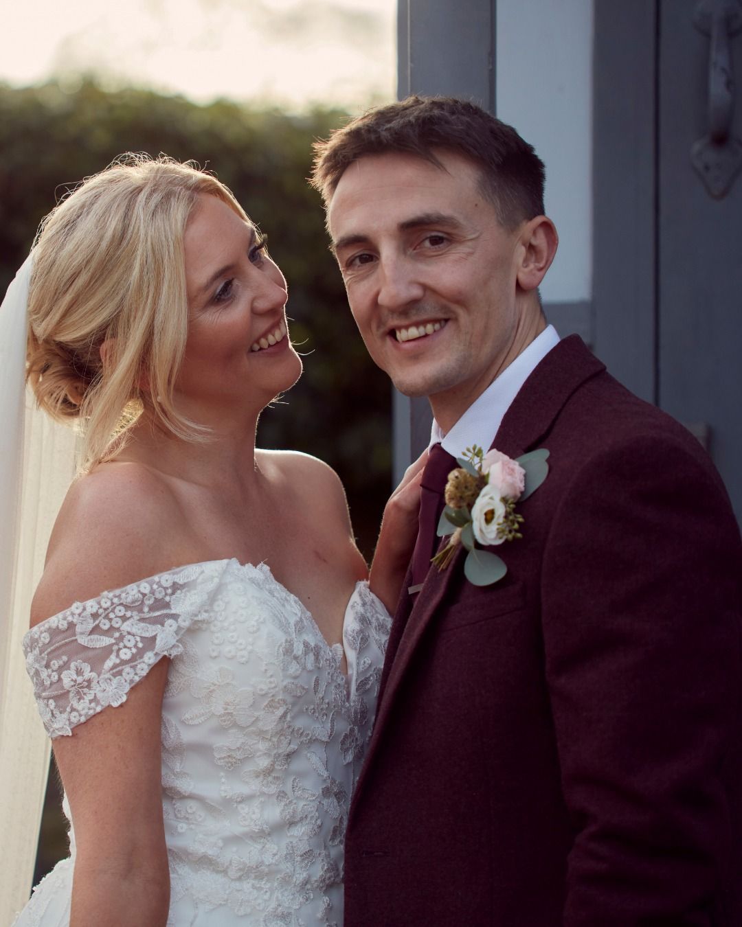 Bride and groom smiling outdoors, bride in white lace dress, groom in burgundy suit with boutonniere