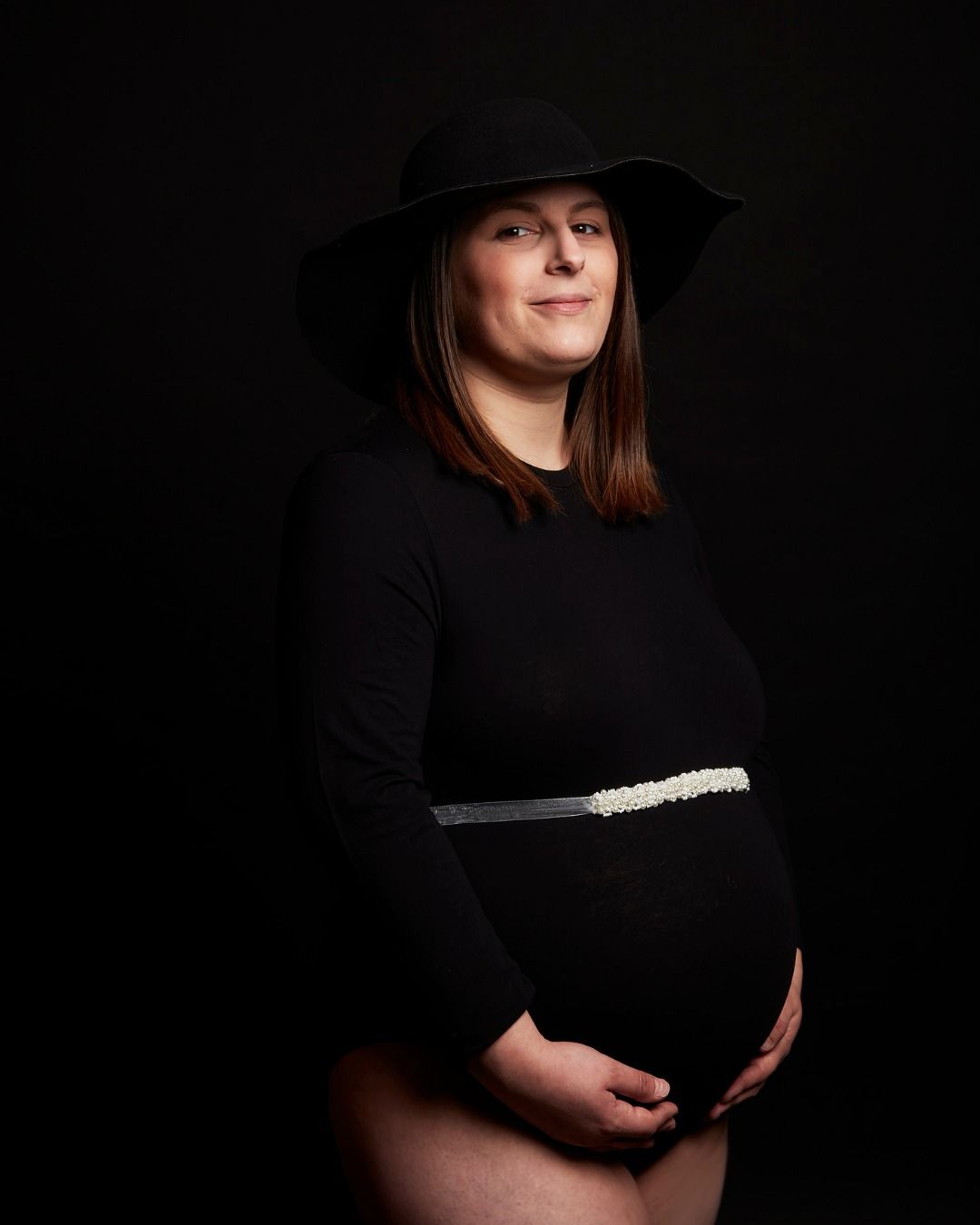 Pregnant person in a black dress and wide-brim hat, standing against a dark background and holding their belly