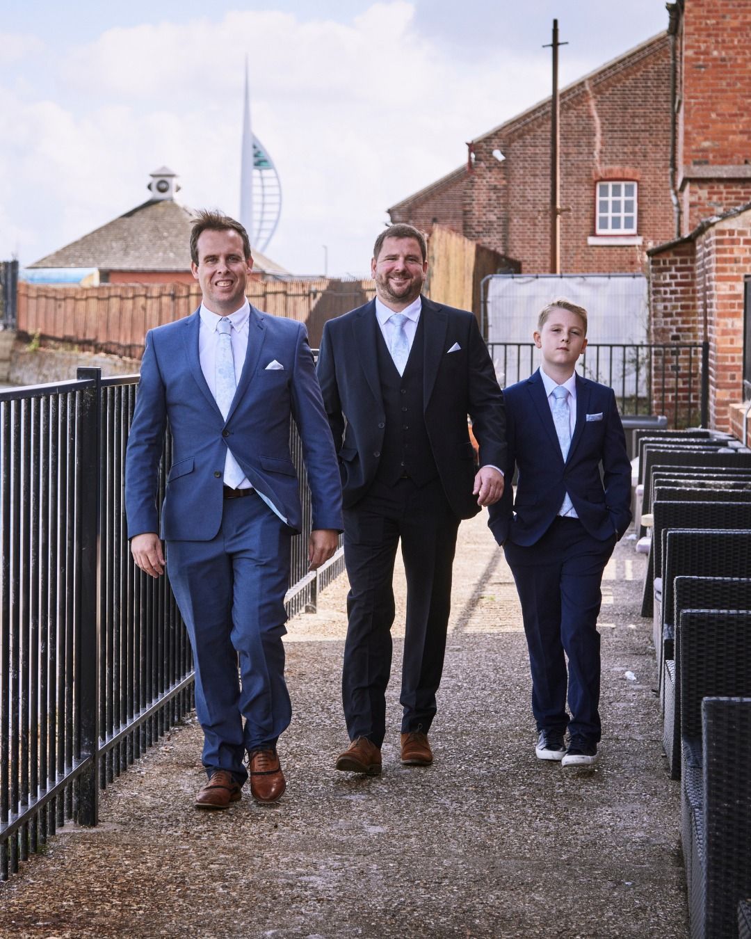 Three men in suits walking down a path beside brick buildings and a railing outdoors