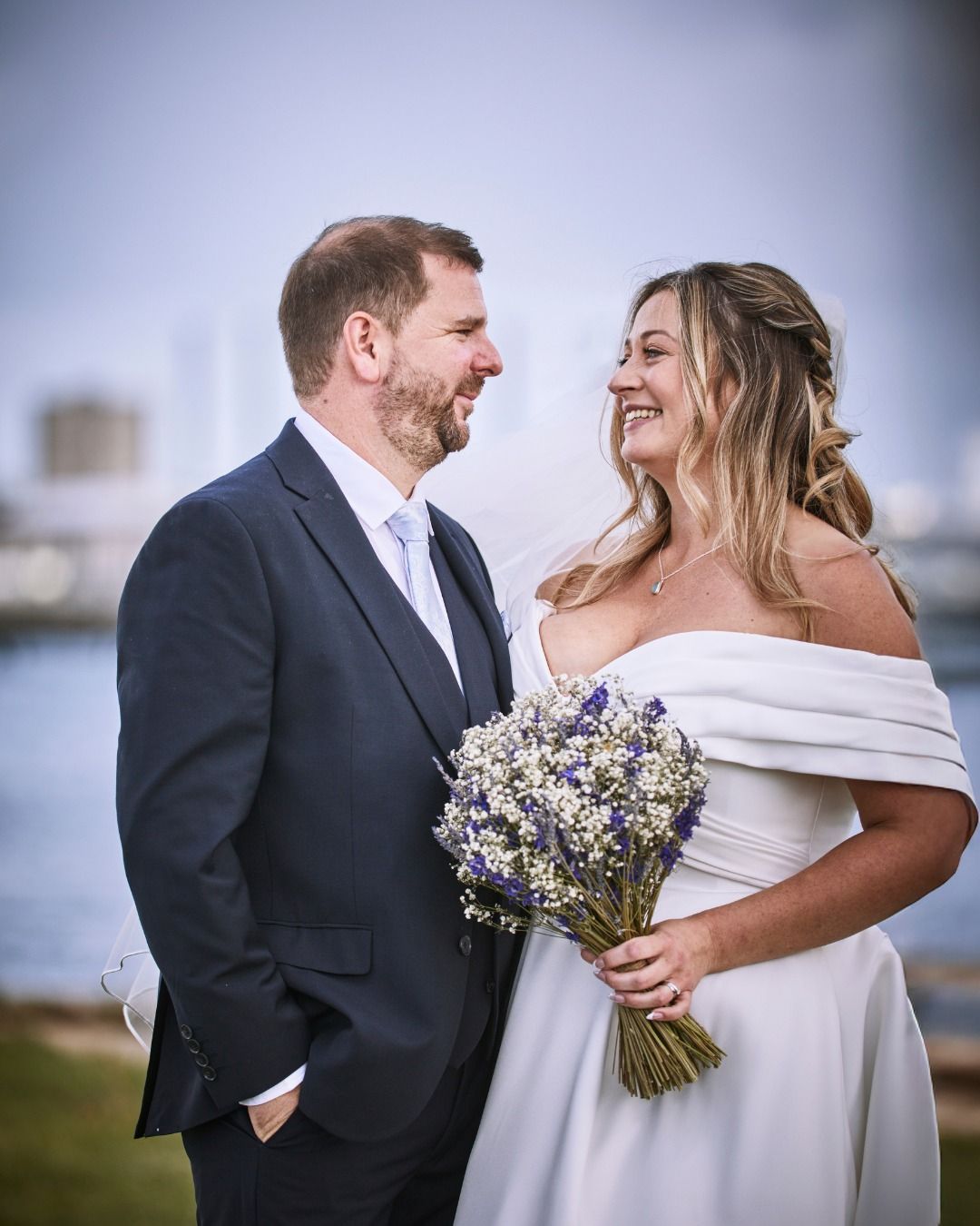 Bride and groom smiling outdoors, the bride holding a bouquet of purple flowers