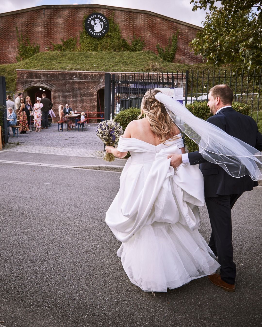 Bride and groom walking toward a rustic brick venue, bride’s veil trailing behind them.