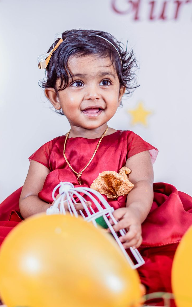Smiling child in a red dress sitting with balloons and a toy basket at a festive backdrop