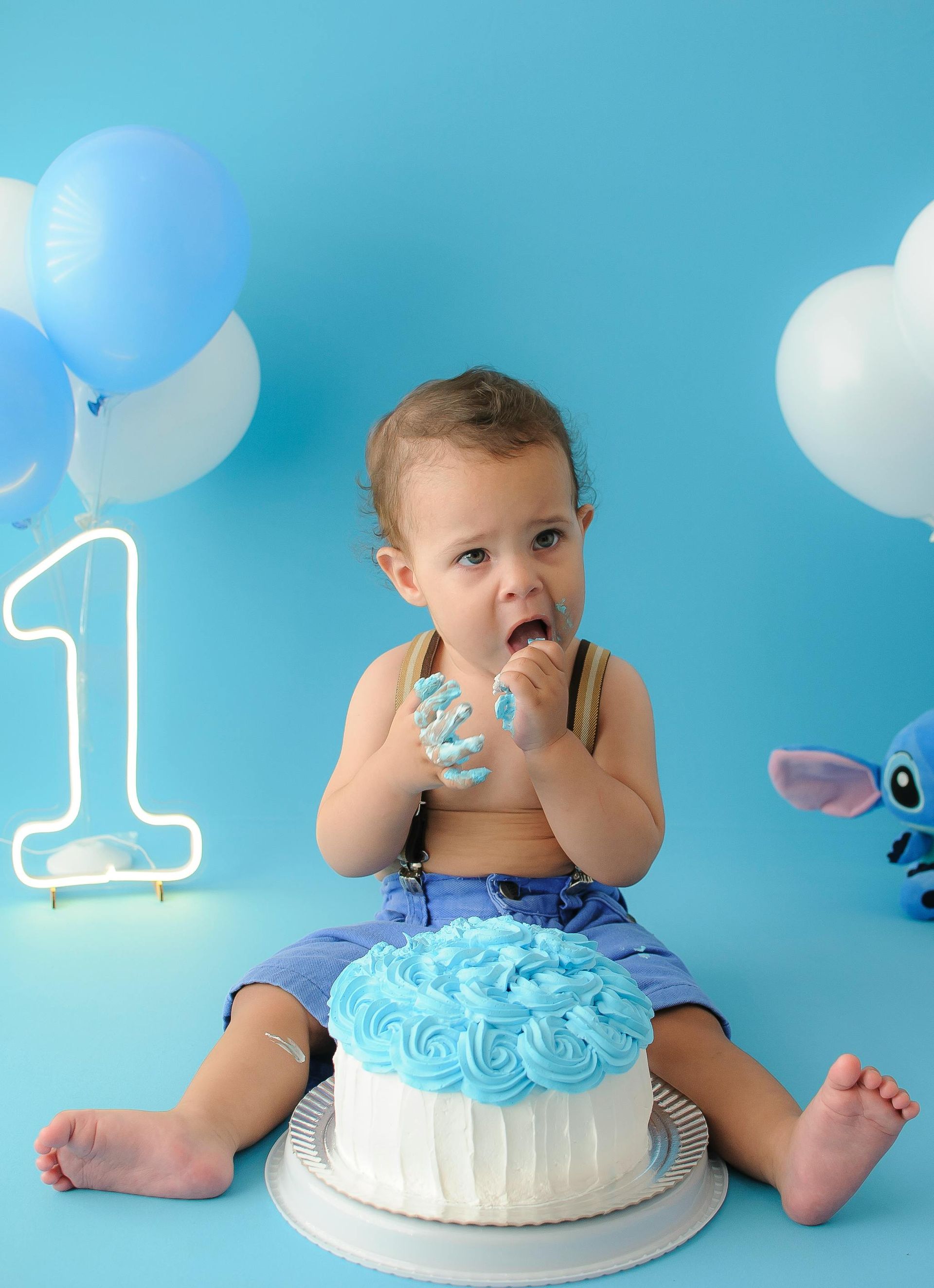 Baby in blue birthday setup, sitting on a cake with balloons and a glowing number 1.