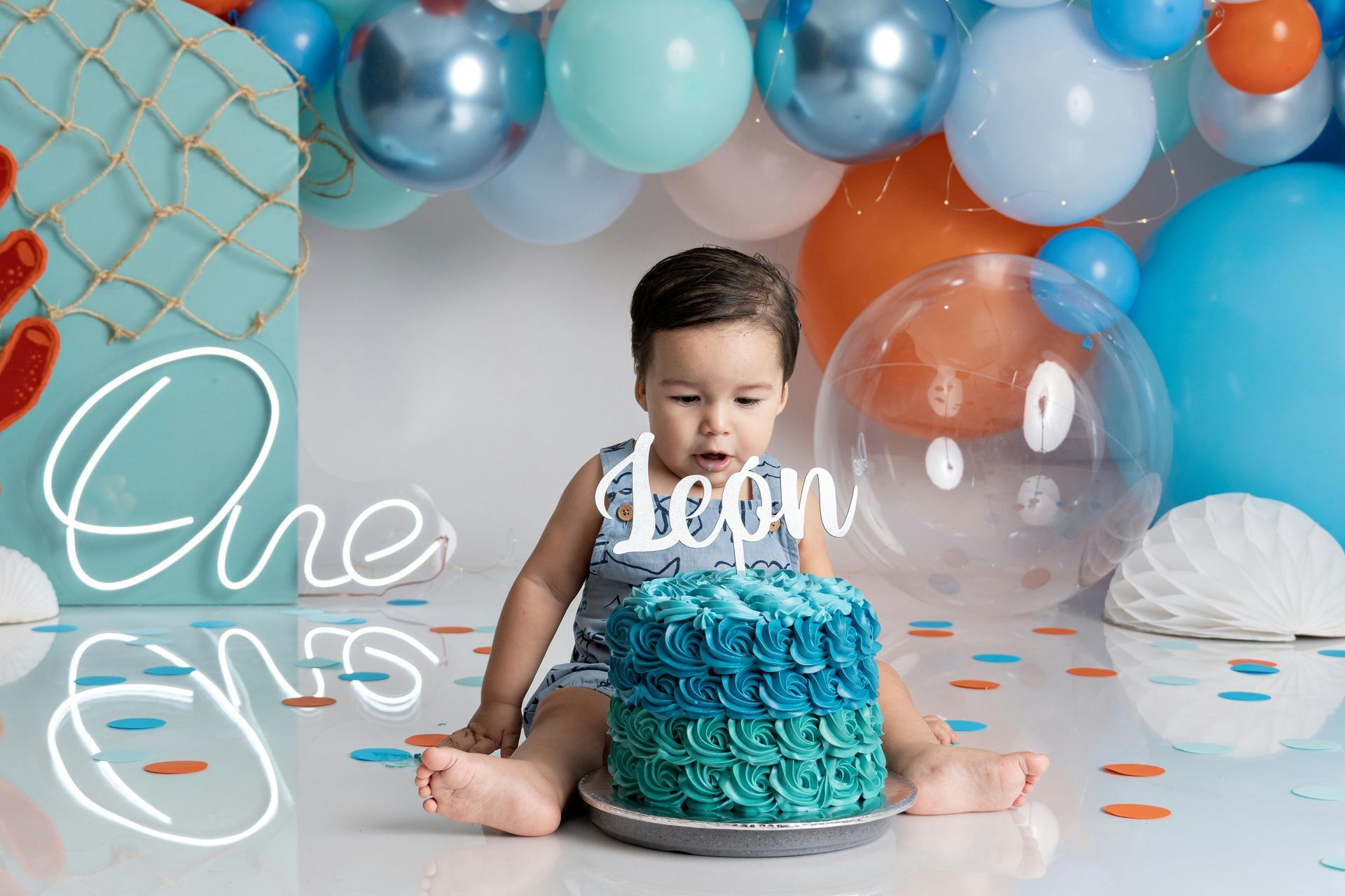 Child sitting by a blue birthday cake with balloon decorations and “One” sign in the background