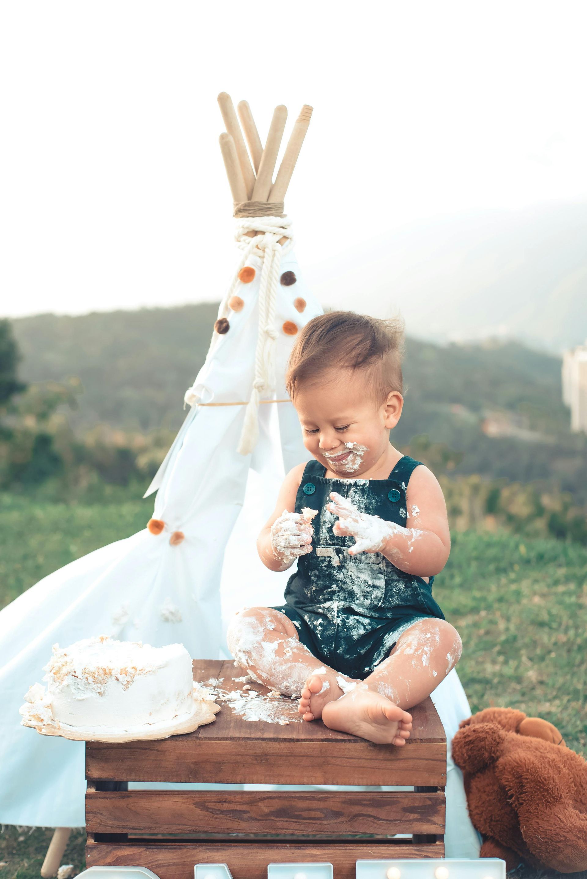 Toddler sitting on wooden crates outdoors, playing with cake beside a small teepee.