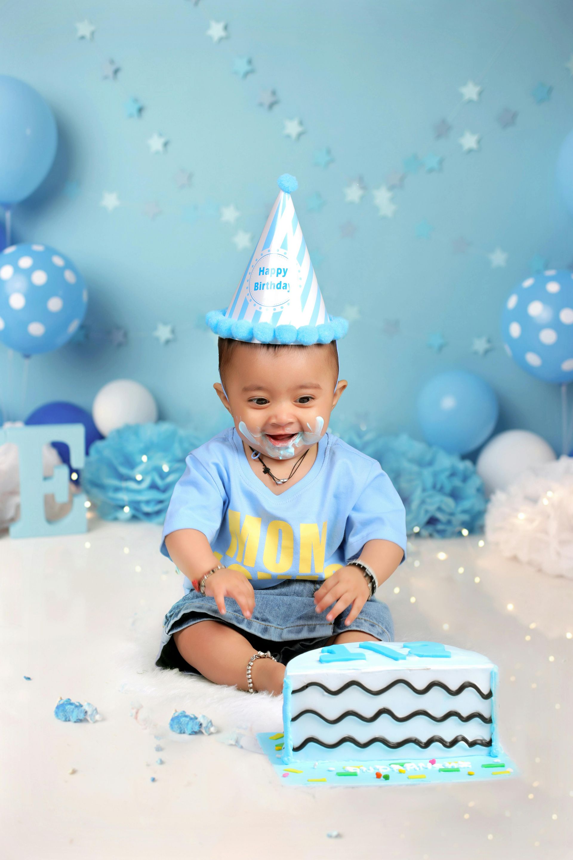 Child in blue party hat smiling at a blue-and-white birthday cake with balloon decorations.