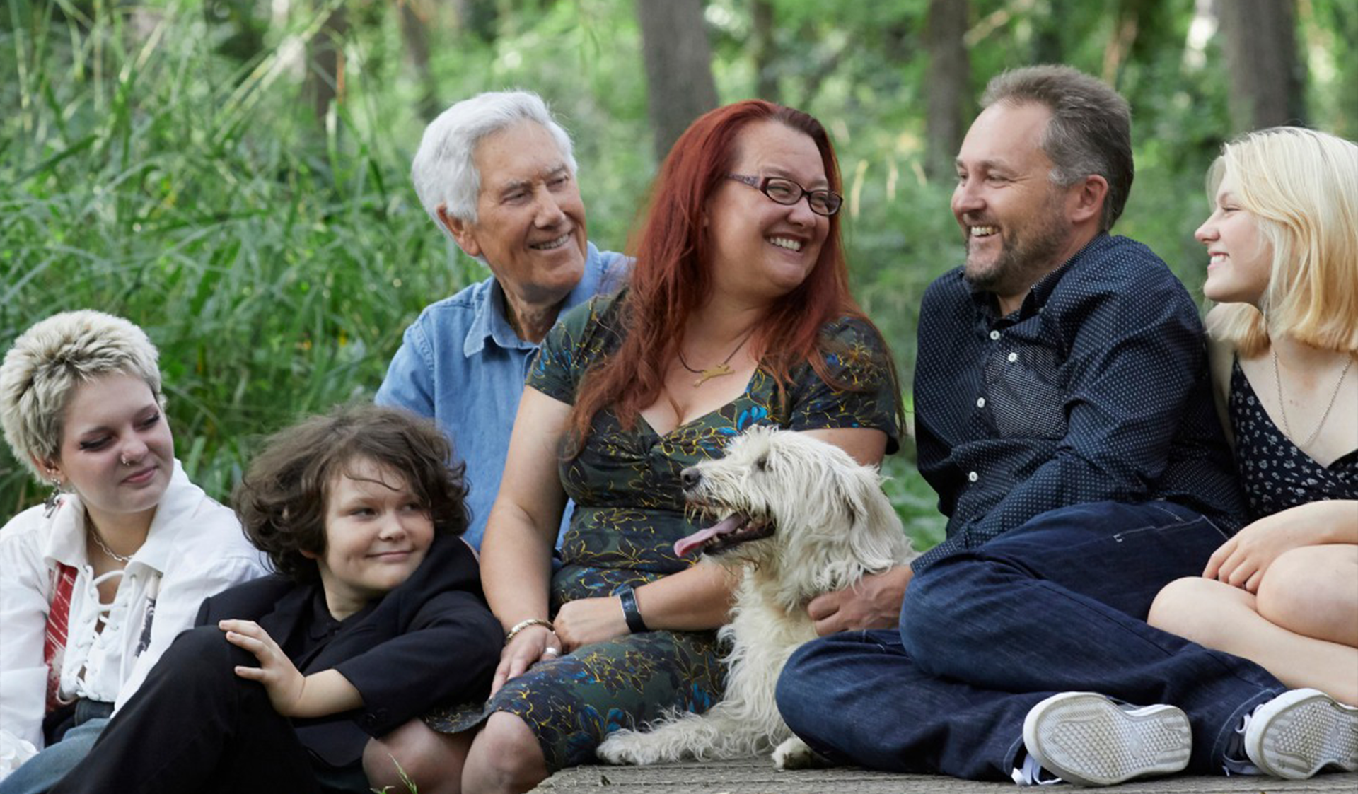 Six people sitting outdoors on grass, smiling and chatting with a small white dog in the center