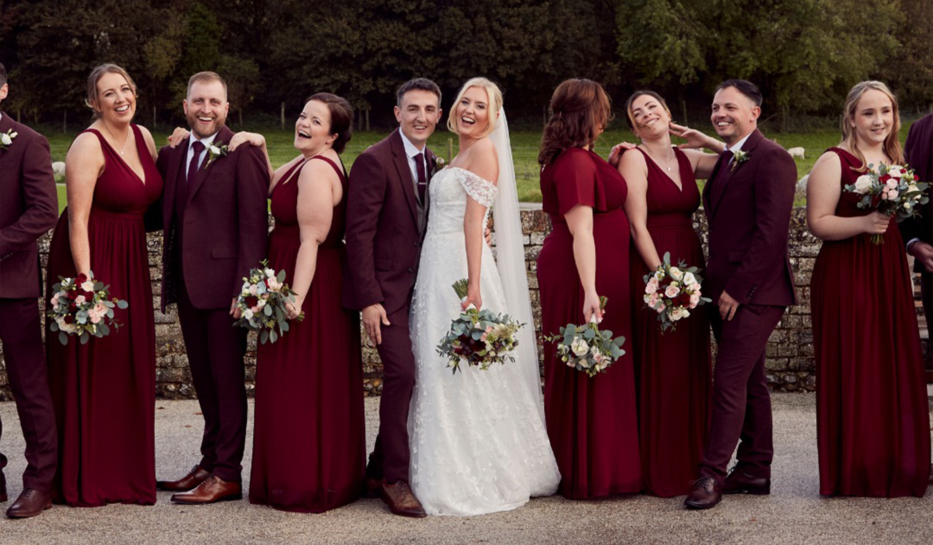 Wedding party in burgundy dresses and suits posing with the bride in a white gown outdoors.