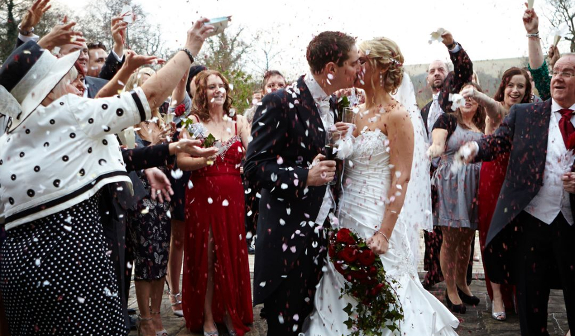 Wedding guests shower a newly married couple with confetti as they kiss outdoors.