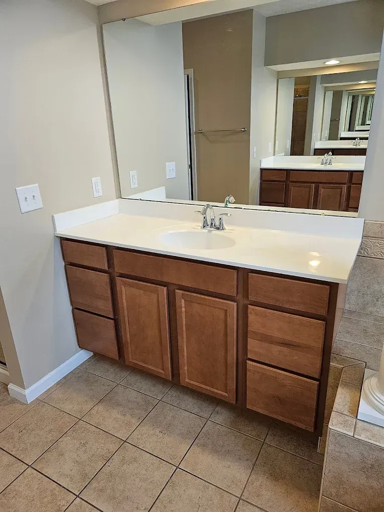Bathroom vanity with light countertop, wooden cabinets, and large mirror reflecting another vanity.
