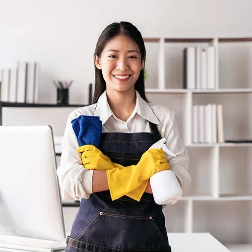 Woman wearing an apron, yellow gloves, and holding a spray bottle and cloth smiles in an office.