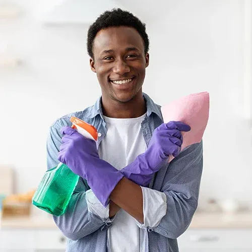 Man smiling, wearing gloves, holding spray bottle and cloth, indoors.