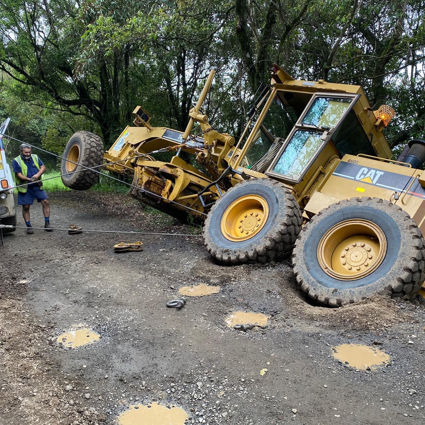 Yellow Caterpillar grader overturned on a muddy road, being towed by a tow truck — Bangalow Towing in Bangalow, NSW