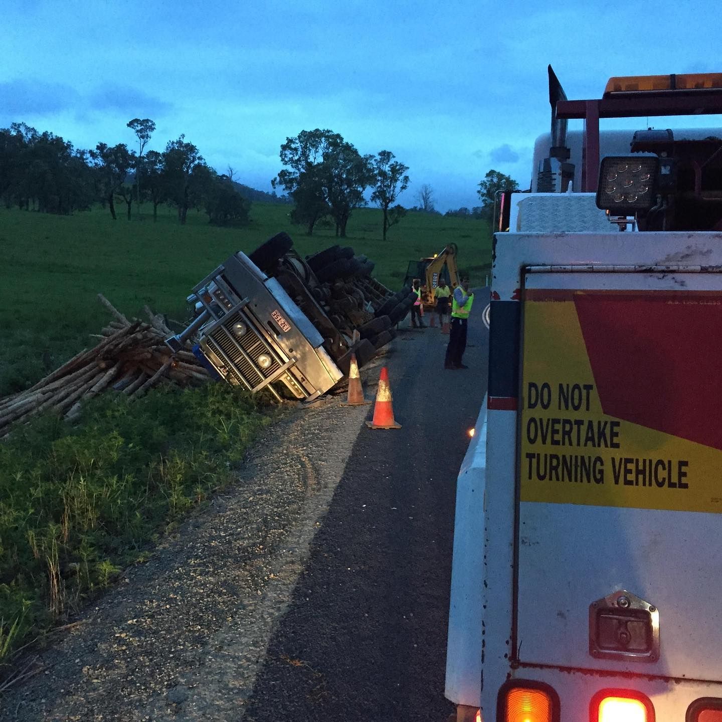 A tipped-over truck on a road; tow truck on right, workers and orange cones on the road — Bangalow Towing in Bangalow, NSW