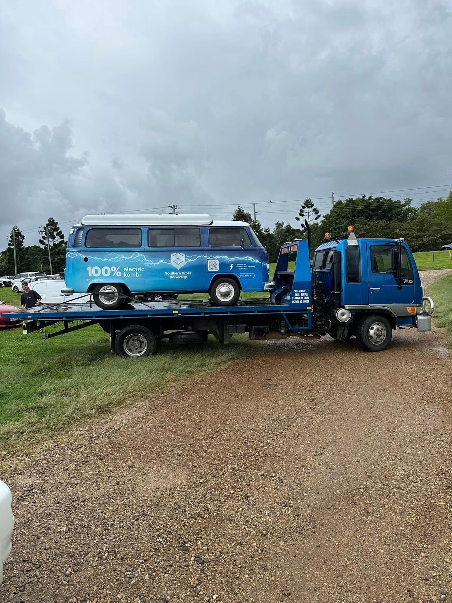 Blue flatbed tow truck carrying a blue van on a gravel road under a cloudy sky — Bangalow Towing in Bangalow, NSW