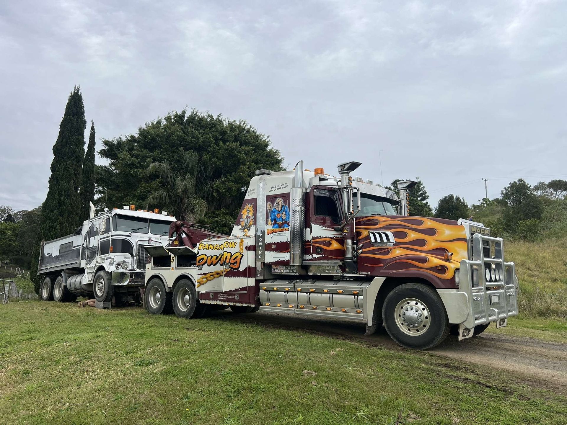 Tow truck hauling a semi-truck on a grassy area; the tow truck has flames painted on its hood — Bangalow Towing in Bangalow, NSW