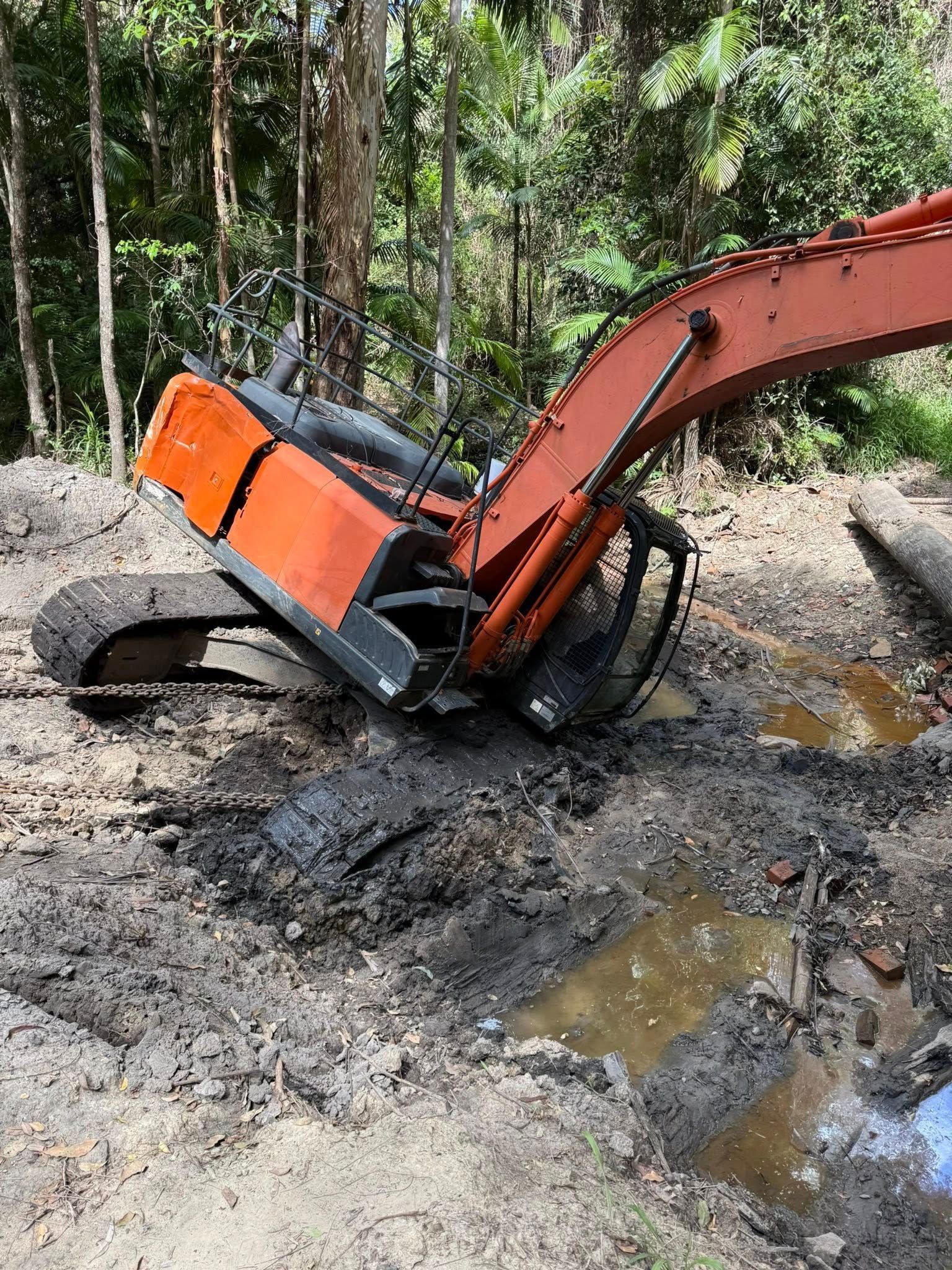 Orange excavator stuck in mud near trees — Bangalow Towing in Bangalow, NSW