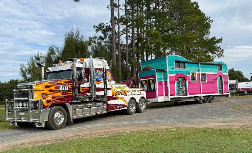 A semi-truck with flame decals pulls a colorful trailer; parked on a gravel road, trees in the background — Bangalow Towing in Bangalow, NSW