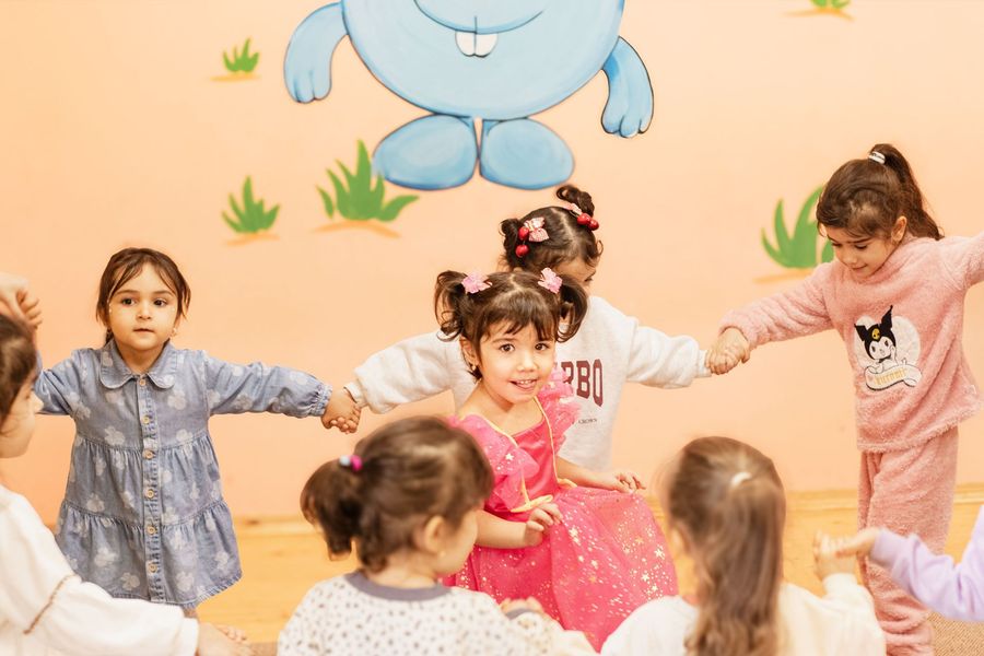 Children holding hands and dancing in a classroom with a large blue cartoon bear on the wall